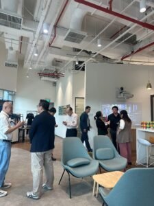 People converse in a modern office with lounge chairs, a projector screen, and snacks on a counter, creating a casual atmosphere